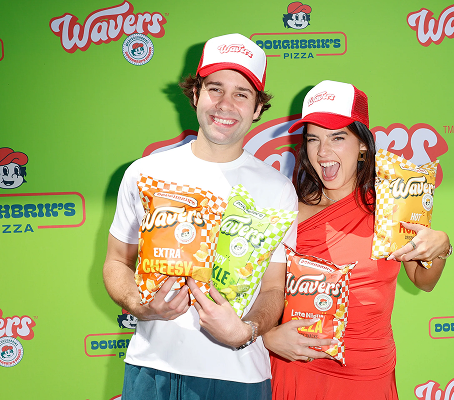 David Dobrik and Natalie Noel pose with Wavers snack bags at product launch event, wearing red and white caps against green branded backdrop
