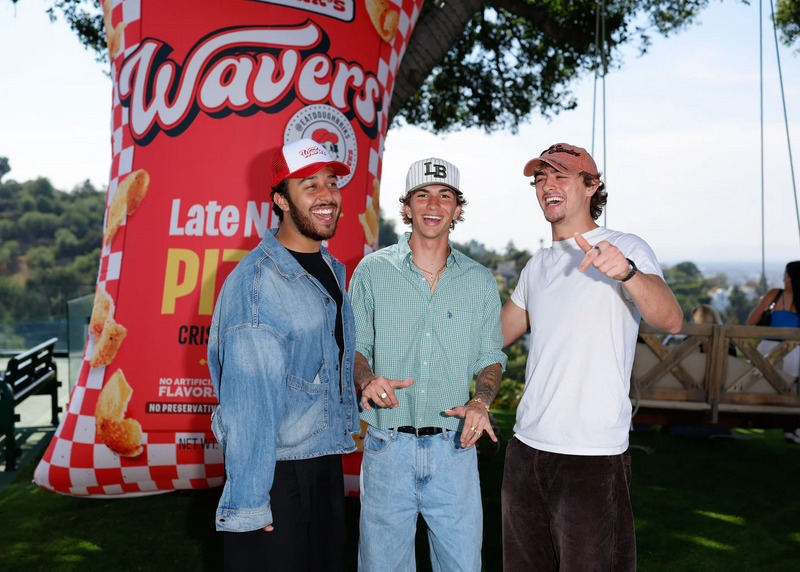 Three young men smiling and posing together outdoors next to a large red Wavy's snack food inflatable advertisement